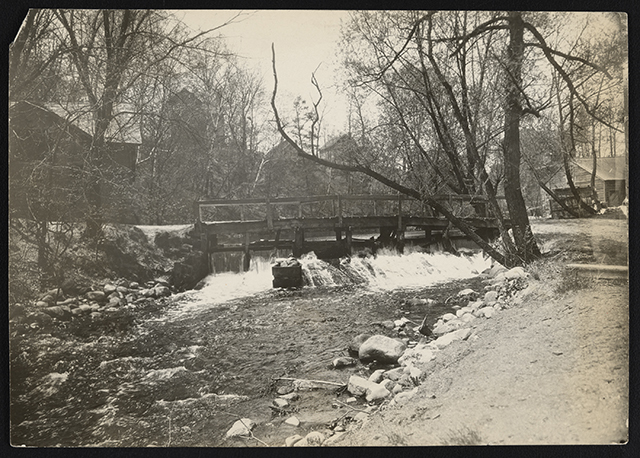 Lightly sepia-toned photograph showing a wooden dam in Bassett Creek. Around the creek and dam are dirt paths, bare trees and wooden houses in the background.